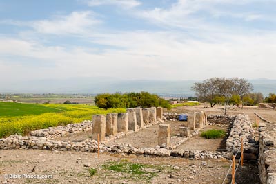 Roughly hewn pillars forming a flanked path through the middle of a structure enclosed with a rectangle of heaped stones, surrounded by dirt and grass overlooked by a blue sky streaked with clouds