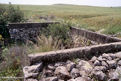 A three sided rectangular stone structure partially overgrown with grasses and shrubs, green hills in the background