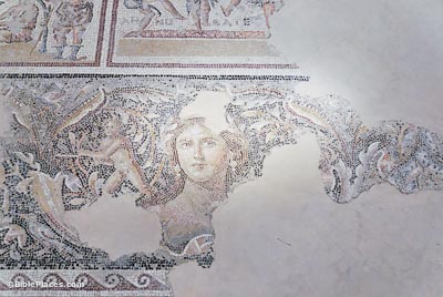 A partially-preserved mosaic on white stone depicting a woman's face surrounded by colorful designs and a winged figure